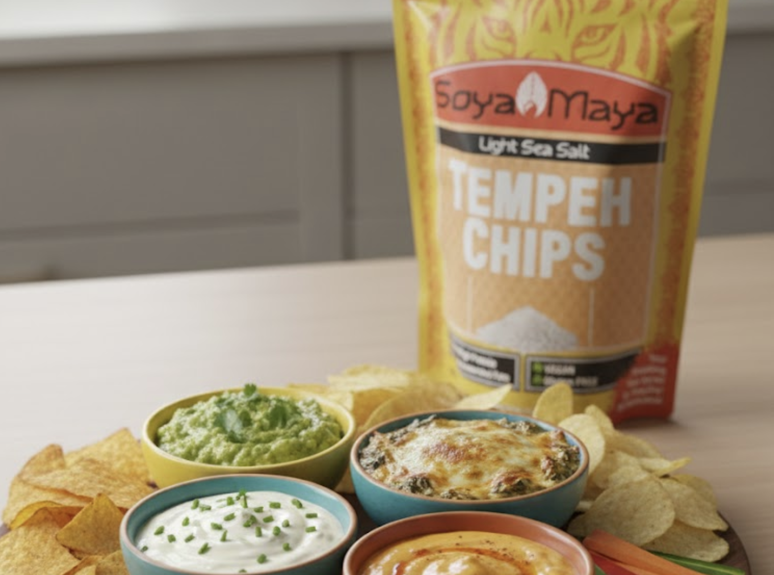 Soya Maya Tempeh Chips with guacamole, sour cream, and cheese dip on a wooden table.