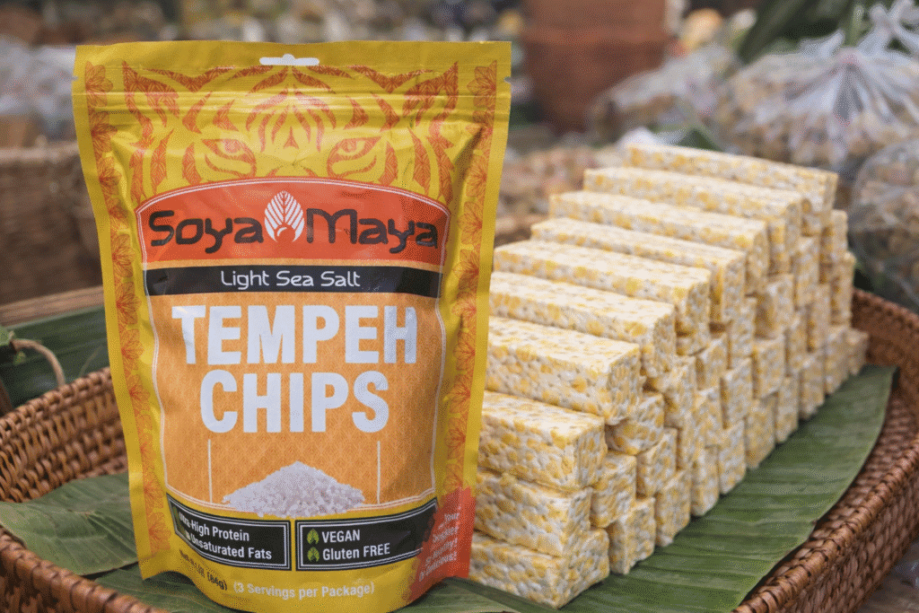 What is Artisanal Tempeh shown with tempeh chips packaging beside stacked fresh tempeh blocks on banana leaves.